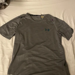 Under armour T-shirt  - Grå under armour T-shirt med reflekterande logga på bröstet och ett riktigt fett mönster över hela tröjan. Helt oanvänd. Tveka inte att höra av dig vid minsta fundering!