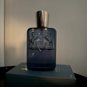 Parfym Sedley från Parfums de Marly - Elegant parfymflaska i mörkblått glas med silverfärgad kork. Flaskan pryds av Parfums de Marlys ikoniska emblem med två hästar. Förpackningen är stilren i matchande blå nyans med silverdetaljer. Perfekt för den som söker en sofistikerad doftupplevelse. Halva kvar typ av 125ml