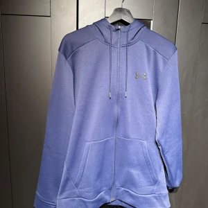 Blå hoodie från Under Armour - Säljer en blå hoodie från Under Armour med dragkedja och justerbar huva. Tröjan har långa ärmar och en praktisk ficka framtill. Perfekt för en avslappnad stil.