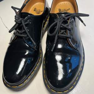 Säljer ett par klassiska svarta Dr. Martens i lack med snygg gul söm runt sulan. Skorna har snörning och är i mycket bra skick. Perfekta för både vardag och fest! Storlek EU 39. Använd en gång. Lite märke på sidan bild fem. Dr Marten 1461 3-hole