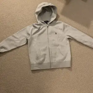 Säljer en stilren grå hoodie från Ralph Lauren i storlek M. Den har en dragkedja framtill och en klassisk logga på bröstet. Perfekt för både vardag och avslappnade tillfällen. Superbekväm och lätt att matcha med allt! 🩶