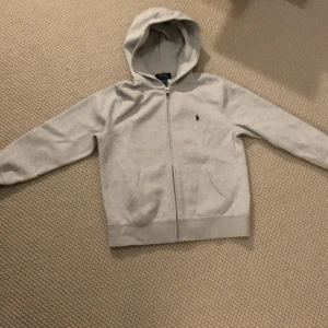 Grå hoodie från Ralph Lauren - Säljer en stilren grå hoodie från Ralph Lauren i storlek M. Den har en dragkedja framtill och en klassisk logga på bröstet. Perfekt för både vardag och avslappnade tillfällen. Superbekväm och lätt att matcha med allt! 🩶