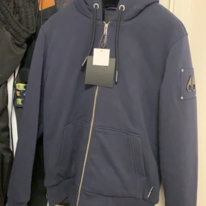 Mörkblå hoodie från Moose Knuckles - Säljer en snygg mörkblå hoodie från Moose Knuckles i nytt skick. Den har en dragkedja framtill och en justerbar huva med snören. På ärmen finns en cool logga. Perfekt för höst och vår, och den är superbekväm att ha på sig. Passar både till vardags och chillkvällar. Kör endast Swish och utanför denna appen, då jag inte vill köra med köparskydd, att det dka gå att lämna tillbaka osv.