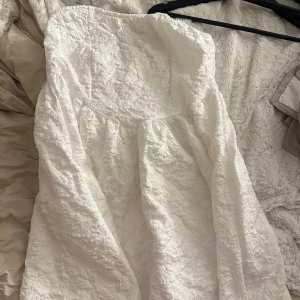 Vit axelbandslös klänning - Säljer en superfin vit axelbandslös klänning i ett härligt blommigt mönster. Klänningen har en luftig passform och är i mycket bra skick. Passar perfekt till studenten! Det står ingen storlek på den men tror att det är L/XL!