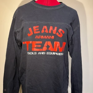 Svart långärmad tröja från Armani Jeans - Cool svart långärmad tröja från Armani Jeans med röd och vit text på framsidan. Perfekt för en avslappnad stil. Tröjan är i bra skick och passar perfekt till jeans eller chinos. Ett måste för garderoben!