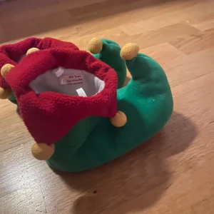 Älvskor - Supermysiga tofflor i grönt och rött med roliga bollar på toppen. Perfekta för att hålla fötterna varma under julen. De är gjorda av mjukt material och har en lekfull design som påminner om tomtens hjälpredor. Perfekta för att sprida lite extra julstämning hemma!