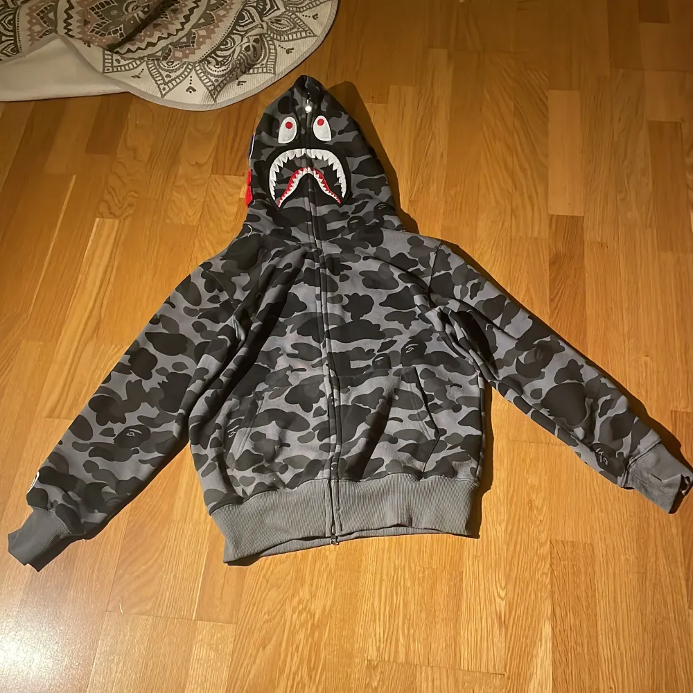 Säljer en grym hoodie från A Bathing Ape i camouflagemönster. Den har en cool hajdesign på huvan och dragkedja framtill. Perfekt för streetwear-stilen och håller dig varm och snygg. Passar perfekt för höst och vår! Skriv vid frågor. Den är M men liten i storlek så typ s. Jackor.