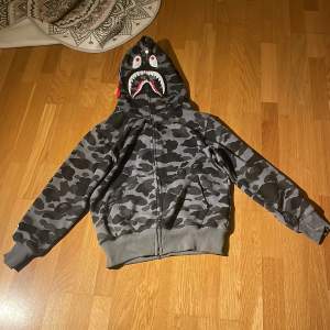 Säljer en grym hoodie från A Bathing Ape i camouflagemönster. Den har en cool hajdesign på huvan och dragkedja framtill. Perfekt för streetwear-stilen och håller dig varm och snygg. Passar perfekt för höst och vår! Skriv vid frågor. Den är M men liten i storlek så typ s