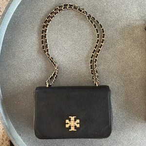 Svart axelväska från Tory Burch - Snygg svart axelväska i skinn från Tory Burch med guldfärgad kedja och logga. Perfekt för både vardag och fest. Väskan har en praktisk dragkedja inuti för säker förvaring. Stilren och tidlös design som passar alla säsonger.