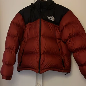 Röd och svart dunjacka från The North Face - Säljer en snygg och varm röd och svart dunjacka från The North Face. Jackan har en dragkedja framtill och är perfekt för kalla vinterdagar. Den är i mycket bra skick och har en normal passform. Perfekt för både vardag och äventyr! Köpt 2022 men knappt någonsin använd!