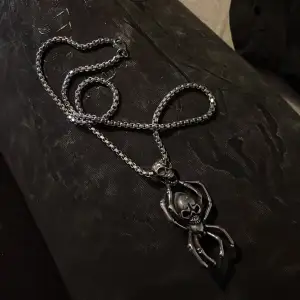 Coolt och edgy halsband i silverfärg med en unik hängande spindel och dödskalle-design. Perfekt för att ge din outfit en tuff touch. Kedjan är robust och hängsmycket är detaljerat med en skrämmande look. Passar perfekt för halloween eller som ett statement-smycke.