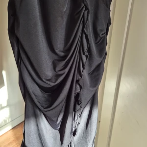 Svart klänning från Vila - Snygg svart klänning från Vila i storlek M. Den har en omlott-design med volangdetaljer längs sidan och ett brett band i midjan för en smickrande passform. Perfekt för festliga tillfällen!