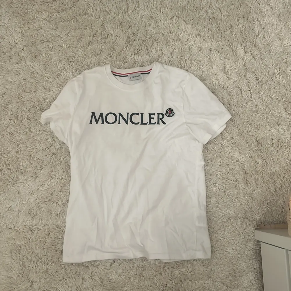 Säljer en snygg vit t-shirt från Moncler. Den har korta ärmar och Moncler-loggan tryckt i svart på bröstet. Perfekt för en stilren och avslappnad look. Passar till både vardag och fest! Inget ingår däremot. Köpt begagnad.. T-shirts.