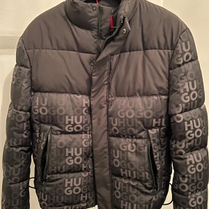 Svart pufferjacka från Hugo - Säljer en snygg svart pufferjacka från Hugo i nytt skick. Jackan har ett coolt mönster med HUGO-loggan över hela ytan. Den är långärmad med dragkedja och knappar framtill. Perfekt för höst och vinter, håller dig varm och stilren. Passar perfekt för både vardag och fest!