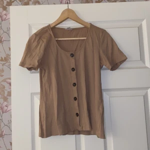 Beige blus från Vero Moda - Säljer en snygg beige blus från Vero Moda i storlek L. Blusen har korta ärmar och knappar framtill. Perfekt för både vardag och festliga tillfällen. Den är i mycket bra skick och redo för en ny ägare!