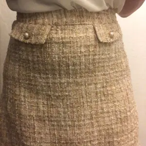 Säljer en superfin beige tweedkjol med stiliga pärldetaljer på fickorna. Kjolen har en klassisk och elegant look, perfekt för både höst och vår. Den är i mycket bra skick och passar perfekt för en chic vardagsstil eller en mer uppklädd look.