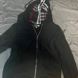 Svart hoodie från Burberry - Säljer en snygg svart hoodie från Burberry med klassiskt rutigt mönster i luvan. Den har en dragkedja framtill och är i mycket bra skick. Perfekt för höst och vår när du vill vara både stilren och bekväm. Passar till alla tillfällen!