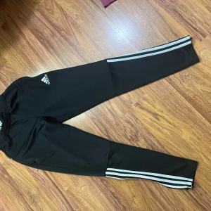 Svarta träningsbyxor från Adidas - Säljer ett par svarta träningsbyxor från Adidas i mycket bra skick. De har de klassiska tre vita ränderna längs sidorna och en dragkedja vid bensluten. Byxorna är tillverkade i ett syntetmaterial och har en elastisk midja för bästa passform. Perfekta för träning eller en avslappnad dag.