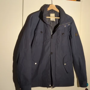 Mörkblå jacka från J.Lindeberg - Säljer en snygg mörkblå jacka från J.Lindeberg i storlek L. Jackan har en stilren design med knappar och dragkedja framtill. Perfekt för höst och vår. passform.Storleken sitter mindre och sitter alltså inte som en vanlig L utan mer som en M. Passar dig perfekt om du är cirka 183 cm. Det finns en defekt och det är att knappen längst ner saknas men det är lätt att laga, det märks inte när man har på sig den. säljer för endast 1500 kr medans nypris är 4400! Priset är ej hugget i sten😃 hör av dig