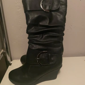 Svarta stövlar med kilklack - Snygga svarta stövlar i skinn med kilklack och dekorativa spännen. Perfekta för höst och vinter, dessa stövlar ger både stil och komfort. De har en slouchy design som ger en avslappnad look. Passar bra till både jeans och klänning.