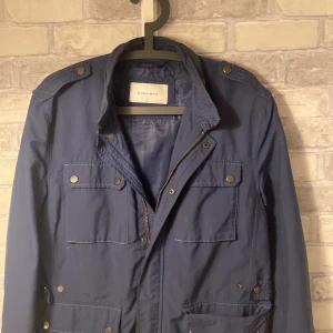 Blå jacka från Zara Man - Säljer en snygg blå jacka från Zara Man. Den har en stilren design med dragkedja och knappar framtill. Perfekt för höst och vår. Jackan har flera praktiska fickor och är i mycket bra skick. Passar både till vardags och lite finare tillfällen.