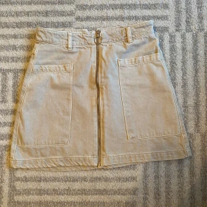 Beige kjol med dragkedja - Säljer en superfin beige kjol i jeansmaterial med en cool dragkedja framtill. Den har två stora fickor fram och bälteshällor i midjan. Perfekt för både vardag och fest! 👜✨