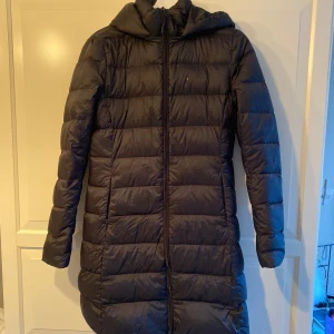 Svart lång dunjacka med huva - Säljer en svart lång dunjacka med huva, perfekt för kalla vinterdagar. Jackan har en dragkedja framtill och är quiltad för extra värme. Den är i mycket bra skick och har långa ärmar. Passar både till vardags och lite finare tillfällen.
