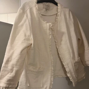Vit blus/kofta från Cream - Säljer en superfin vit blus/kofta från Cream i storlek M. Den har en elegant struktur med volangdetaljer längs kanterna och två praktiska fickor framtill.