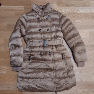 Dunjacka - Säljer en superfin beige dunjacka från Yes! Do Kid's i storlek 12, den sitter som Xxs/xs för damer. Jackan har en mysig pälsfodrad insida och är perfekt för kalla vinterdagar. Den har långa ärmar, dubbla dragkedjor på insidan och knappar framtill samt ett bälte i midjan för extra stil. Perfekt för både vardag och festliga tillfällen! Jackan har ingen pris lapp kvar då den är använd endast en gång och är i helt nytt skick.