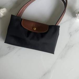 Snygg och praktisk svart axelväska från Longchamp med bruna skinnhandtag och lock. Väskan har en knappdetalj i guld och är perfekt för både vardag och fest. Den är rymlig och stilren, vilket gör den till ett måste i garderoben.Storlek Large