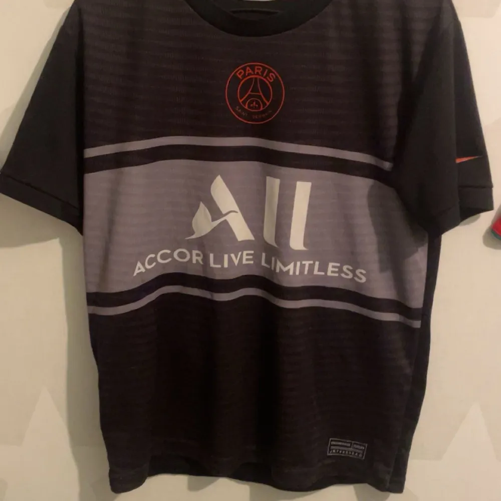 Säljer en svart PSG fotbollströja med Hakimi och nummer 2 på ryggen. Tröjan har korta ärmar och är i bra skick. Den har en röd Nike-logga på ärmen och texten 'Accor Live Limitless' på framsidan. Perfekt för alla PSG-fans!. T-shirts.