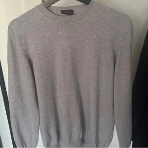 Grå tröja i merinoull från Oscar Jacobson - Säljer en stilren och bekväm grå tröja från Oscar Jacobson i 100% merinoull. Perfekt för kyliga dagar med sin mjuka och värmande kvalitet. Tröjan har en klassisk passform och långa ärmar. Ett måste för garderoben! 🐑