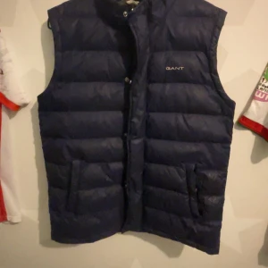 Mörkblå dunväst från GANT - Säljer en snygg mörkblå dunväst från GANT i mycket bra skick. Västen har en quiltad design och stängs med dragkedja och knappar. Perfekt för höst och vår när du vill hålla dig varm utan att bära en jacka. Passar ungdomar i åldern 13-14 år.kan mötas ta inte handen Skärholmen 