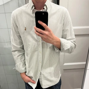 Randig skjorta från Ralph Lauren - Säljer en stilren och klassisk vit och grön randig skjorta från Ralph Lauren. Skjortan har långa ärmar och en liten broderad logga på bröstet. Använd endast 1 gång! Perfekt för både vardag och fest!💚 Passar bra till jeans eller chinos för en avslappnad men ändå uppklädd look.