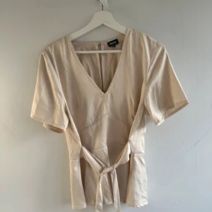 Beige blus med knytband - Snygg beige blus från bubbleroom i storlek M. Den har en v-ringad hals och korta ärmar. Blusen är i ett mjukt material och har ett knytband i midjan för en fin passform. Perfekt för både vardag och fest!