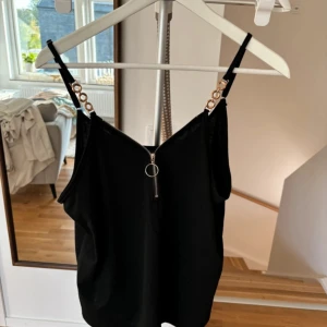 Svart linne med kedjedetaljer - Snyggt svart linne med tunna axelband och coola kedjedetaljer i guld. Linnet har en dragkedja framtill som ger en edgy look. Perfekt för en utekväll eller fest! 🖤✨