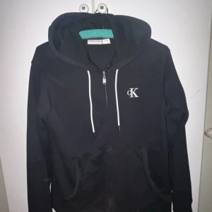 Svart hoodie från Calvin Klein - Säljer en snygg svart hoodie från Calvin Klein i storlek S. Den har en dragkedja framtill och en klassisk CK-logga på bröstet. 
