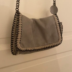 Grå axelväska med kedja - En praktisk och lite mindre handväska i mocka med supersnygga kedjedetaljer som liknar Stella McCartney väskorna. Köpt här på plick men har inte används supermycket, så skicket är fortfarande gott!💕