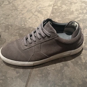 Grå sneakers i mocka - Säljer ett par snygga grå sneakers i mocka. De har en klassisk design med snörning och en vit sula som ger en stilren look. Perfekta för både vardag och lite mer avslappnade tillfällen. Skorna är i bra skick och redo för nya äventyr!     Storlek, 43 från arigato pris kan diskuteras vid snabb affär 