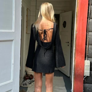 Svart glittrig klänning med öppen rygg - Snygg svart klänning med glittrigt material och öppen rygg. Den har långa ärmar och en tight passform. Klänningen knyts i ryggen för en extra detalj. Perfekt till nyår. Endast använd en gång.