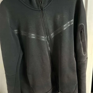 Svart hoodie från Nike - Säljer en svart hoodie från Nike i bra skick. Den har en dragkedja framtill och en praktisk ficka med dragkedja på ärmen. Perfekt för höst och vår, med en stilren design och bekväm passform. Huvan ger extra värme och stil. Passar både till träning och vardag.