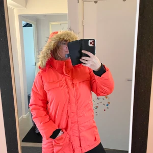 Orange dunjacka med pälshuva - Säljer en snygg och  orange-röda dunjacka med en mysig pälshuva. Jackan har långa ärmar och praktiska fickor framtill. Perfekt! 🧡Helt ny