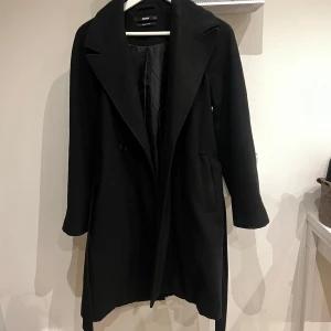 Svart kappa - Säljer en stilren svart kappa från BikBok. Perfekt nu inför kallare väder. Det enda är att sömmen till ena fickan har öppnats upp lite (framgår i bild). 
