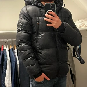 Svart pufferjacka från Zavetti Canada - Säljer en svart pufferjacka från Zavetti Canada i mycket bra skick. Jackan har en snygg logga på ärmen och dragkedja framtill. Perfekt för kalla vinterdagar med sin varma och vadderade design. Den har en normal passform och är superbekväm att ha på sig. Passar perfekt till både vardag och lite mer uppklädda tillfällen.