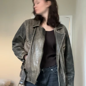 Grå skinnjacka med bälte och dragkedjor - Snygg grå/brun skinnjacka med dragkedja och bälte i midjan. Jackan har en cool vintage-look och två fickor med dragkedja framtill. Den är rejäl i materialet och passar perfekt till en edgy stil. Förutom dragkedjan som lagats med ett litet snöre så är varan i toppenskick tack vare det hållbara materialet! 