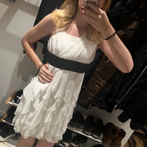 Vit klänning med svart band - Elegant vit klänning med ett svart band i midjan och en dekorativ rosett baktill. Klänningen har en omlott-design med volanger och är ärmlös, vilket ger en luftig och stilren look.