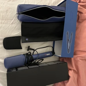 Blå plattång från ghd - Säljer en blå ghd Duet Style plattång med tillhörande värmeskydd och förvaringsväska. Perfekt för att skapa släta och glansiga frisyrer. Kommer i originalförpackning. NYPRIS 4 399kr