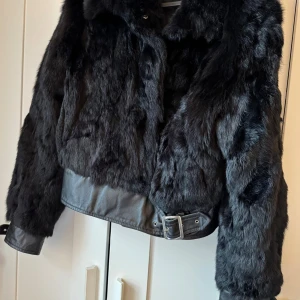 Svart pälsjacka med bälte - Snygg svart pälsjacka med bälte i midjan. Jackan har en lyxig look med långa ärmar och en mjuk pälsstruktur. Perfekt för nu.