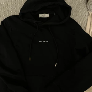 Svart hoodie från Les Deux M - Snygg svart hoodie från Les Deux med justerbar huva och dragsko. Perfekt för en avslappnad stil. Den har en enkel design med märkeslogga på bröstet.