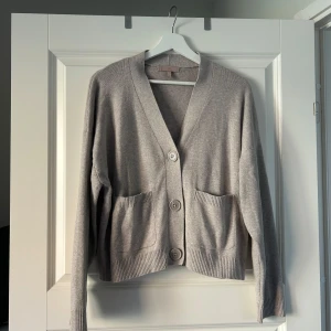 Beige kofta med knappar - Säljer en stilren beige kofta med stora knappar framtill och praktiska fickor. Koftan har långa ärmar och ribbade kanter som ger en klassisk look. Perfekt för kyligare dagar.